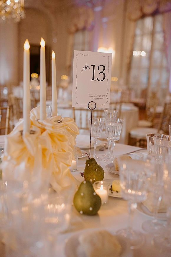 Reception tablescape with a wedding table number card, taper and votive candles, ribbon bow centerpiece, pears, and white linens in a chandelier-lit room