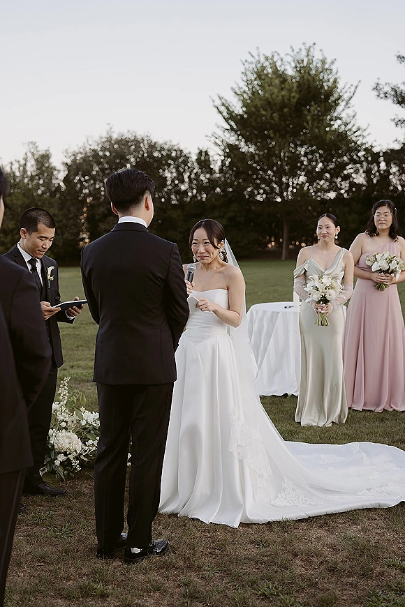 Wedding vows as bride reads into a microphone facing groom in black tuxedo, veil and bouquet on an outdoor lawn with trees and sky