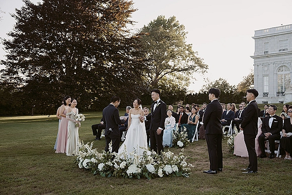 Ceremony moment at an outdoor wedding ceremony as bride and groom stand at a floral arch with wedding party, guests on lawn by mansion