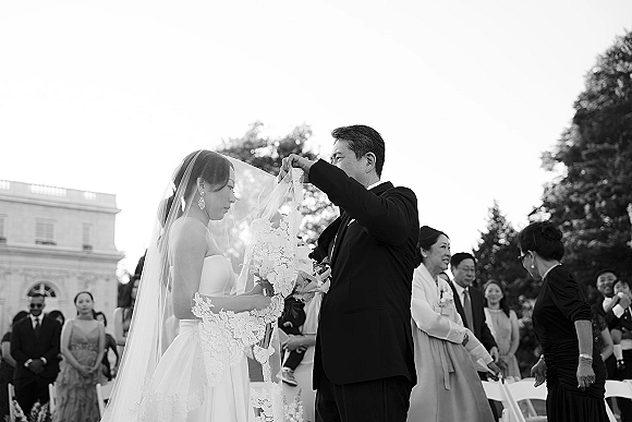 Ceremony moment at an outdoor wedding ceremony as the groom lifts the bride’s veil, she holds a bouquet, guests watch on the lawn