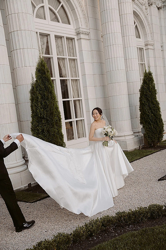 Bride portrait in a strapless satin wedding dress holding a white calla lily bouquet as her cathedral veil flows by stone columns