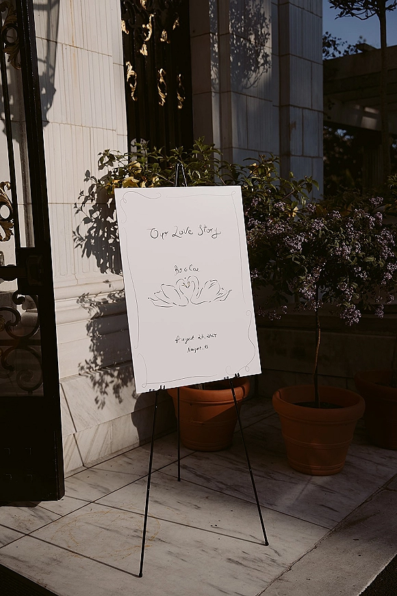 Wedding welcome sign on an easel with hand-drawn lettering and floral illustration, set by a stone doorway with potted plants
