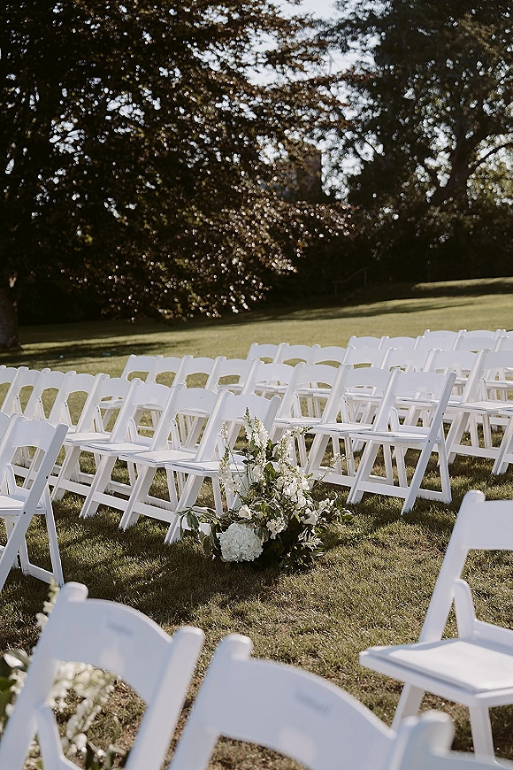 Outdoor ceremony seating with wedding aisle chairs in neat rows, white folding chairs and a white-and-green floral aisle marker on a sunlit lawn