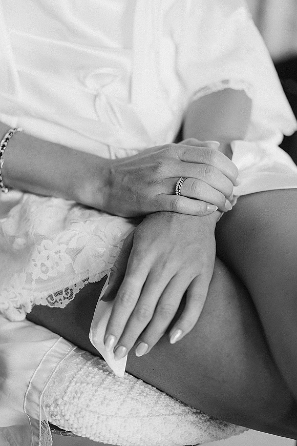 Wedding rings close-up on bride’s manicured hands, diamond wedding band stacked beside a bracelet and lace robe over upholstered lap