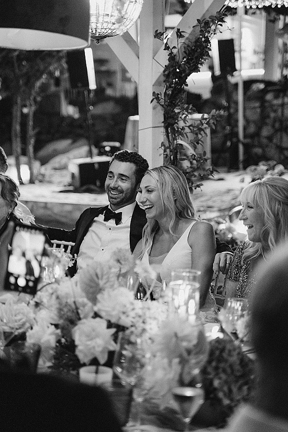Reception moment as bride and groom laughing at their head table, candlelit florals and glassware under string lights on an outdoor patio