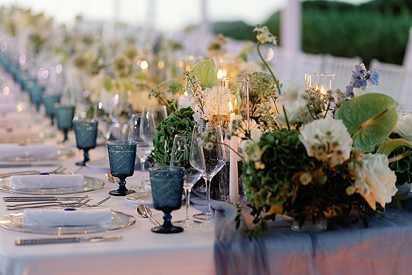 Reception tablescape with wedding table centerpiece of white flowers and blue hydrangea, tall candles and blue goblets on an outdoor long table