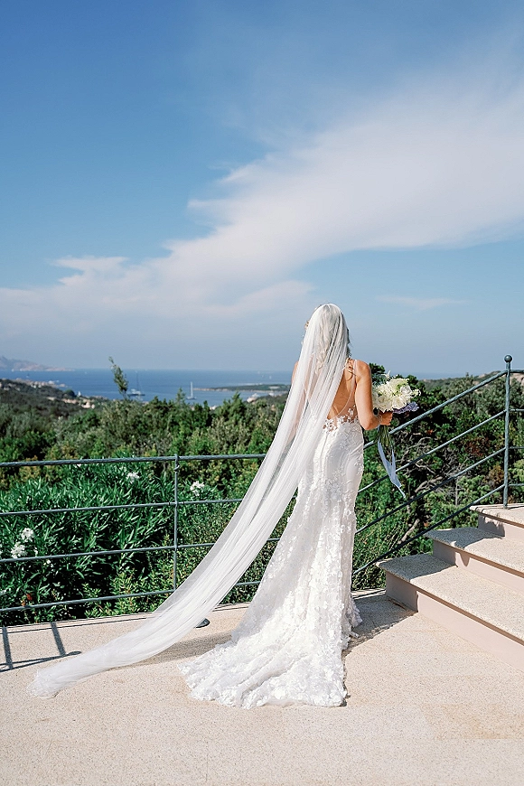 Bridal portrait of a back view bride in a lace mermaid gown with long cathedral veil, holding a bouquet on a seaside terrace railing