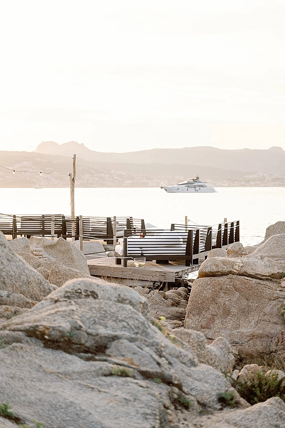 Waterfront lounge setup with outdoor sofa seating and striped cushions on a wooden deck under string lights, overlooking rocky shore and ocean