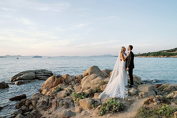 Couple portrait of bride and groom by ocean, embracing on rocky coastline as her veil and dress trail in the wind, boats behind