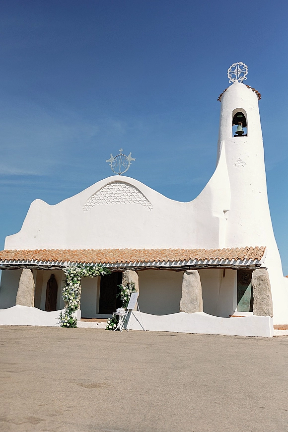 Chapel wedding entrance framed by a church entrance floral arch of white and green flowers, with welcome sign in a sunny courtyard under blue sky