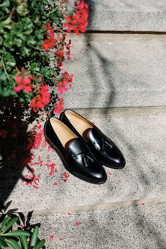 Groom shoes, black tassel loafers resting on stone steps with flower petals and greenery, sunlight casting soft shadows around them
