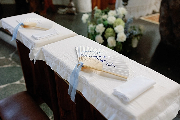 Wedding ceremony program with ceremony program fans tied in blue ribbon bows on a linen runner, set on wooden benches near florals indoors