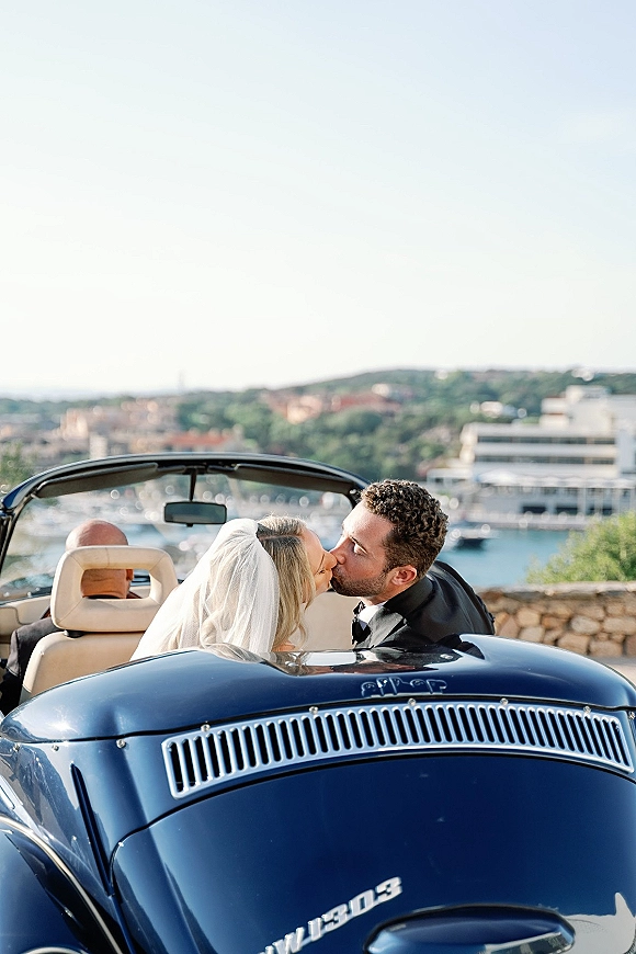 Wedding kiss as bride and groom in car, her veil flowing beside his tuxedo in a vintage convertible by a harbor overlook under blue sky
