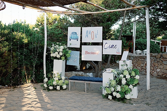 Wedding photo booth with hanging backdrop signs, string lights, and white floral pedestals beside a bench on sandy ground by trees