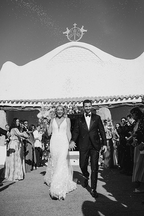Wedding recessional as newlyweds walking aisle through confetti, bride in lace dress with bouquet and veil beside tuxedoed groom outside chapel facade