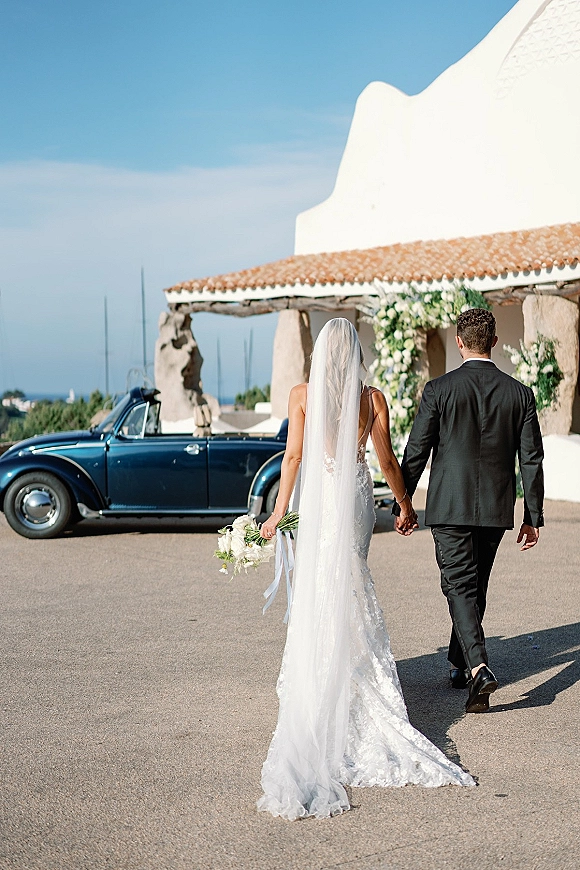 Wedding couple portrait of newlyweds holding hands walking away, bride’s long veil and calla lily bouquet beside a vintage convertible car