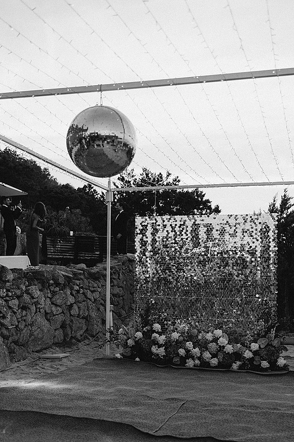 Reception dance floor with mirror disco ball decor under a string light canopy, sequin wall and neon sign on an outdoor patio by trees