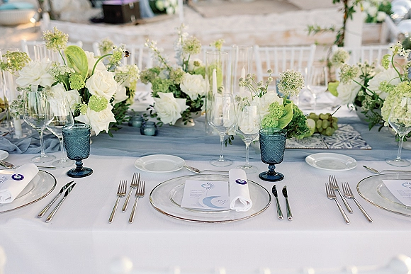 Reception tablescape with wedding table centerpiece of white and green florals, blue goblets, taper candles, and grapes on a blue runner in a blurred reception space