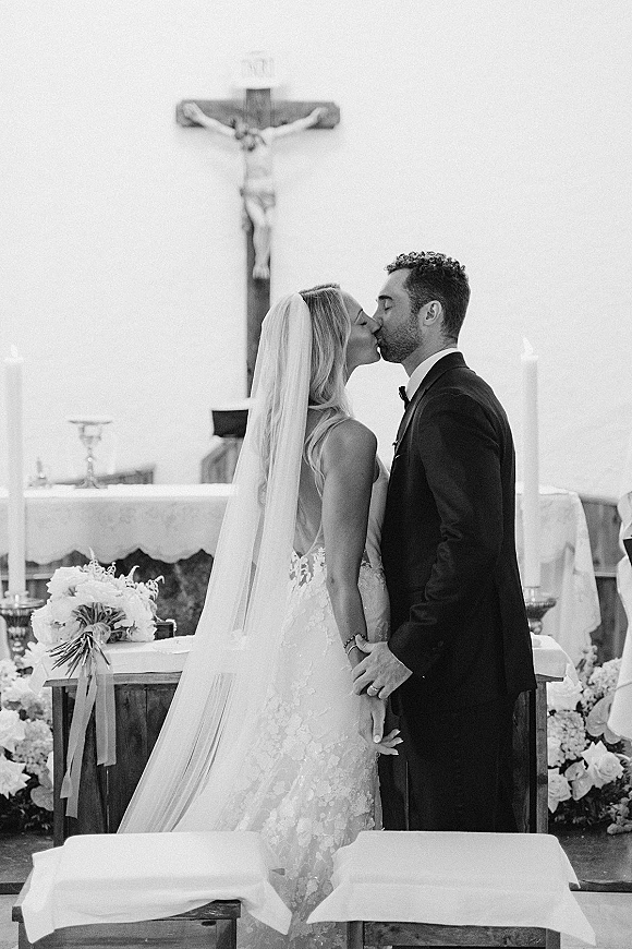 Wedding kiss at a church altar as bride in lace gown and veil kisses groom in tux, with candles, flowers, and crucifix behind