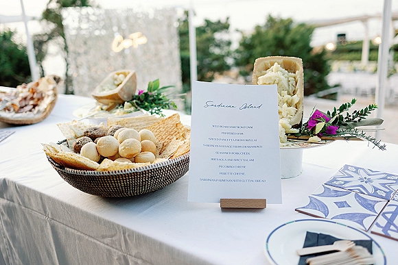 Wedding menu card on a wooden holder beside a charcuterie spread with purple flower accent on a white linen table under a pergola