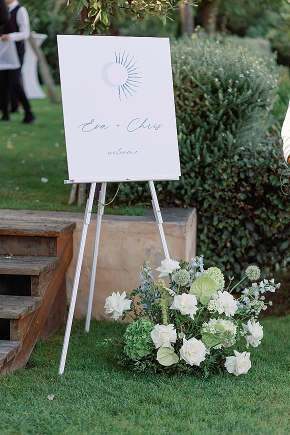 Wedding welcome sign on easel with white roses and green hydrangea at the base along a garden walkway with shrubs and trees