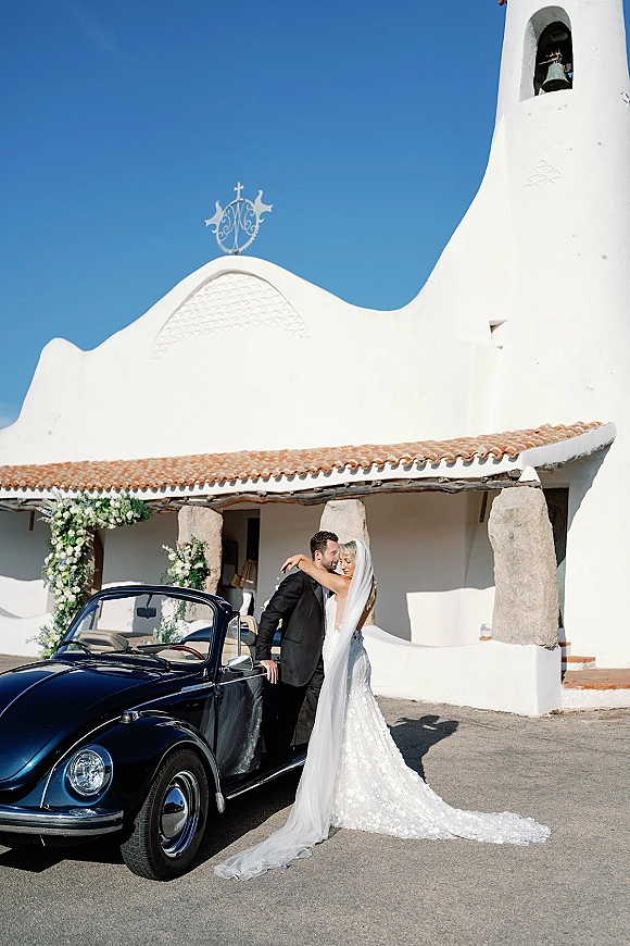 Couple portrait of bride and groom by car, her long veil and lace train beside a vintage convertible outside a white chapel bell tower.