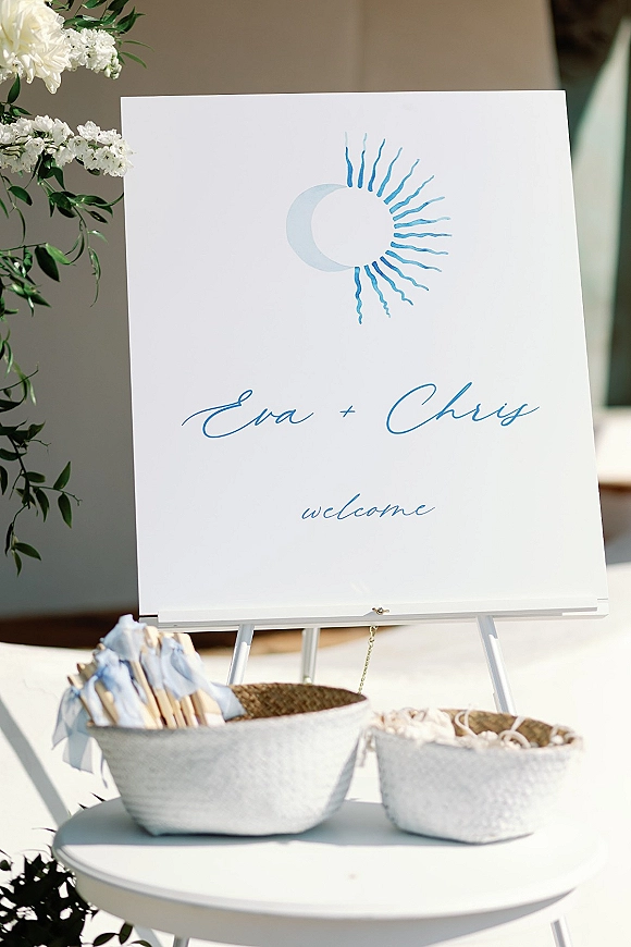 Wedding welcome sign with calligraphy welcome sign in blue on an easel, sun and moon icon, floral greenery, and ribbon wand baskets in sunlight