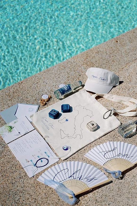 Wedding flat lay with wedding invitation suite, rings, ribbon bows, fans, tote and cap arranged on a stone pool deck by blue water