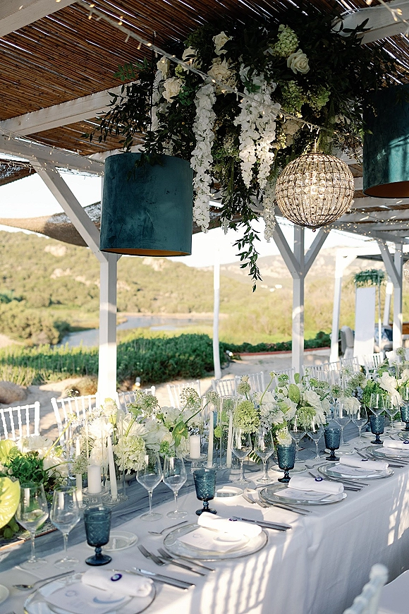 Reception tablescape with long head table decor, white florals and greenery garland, candles and blue goblets under chandeliers on a patio with water view