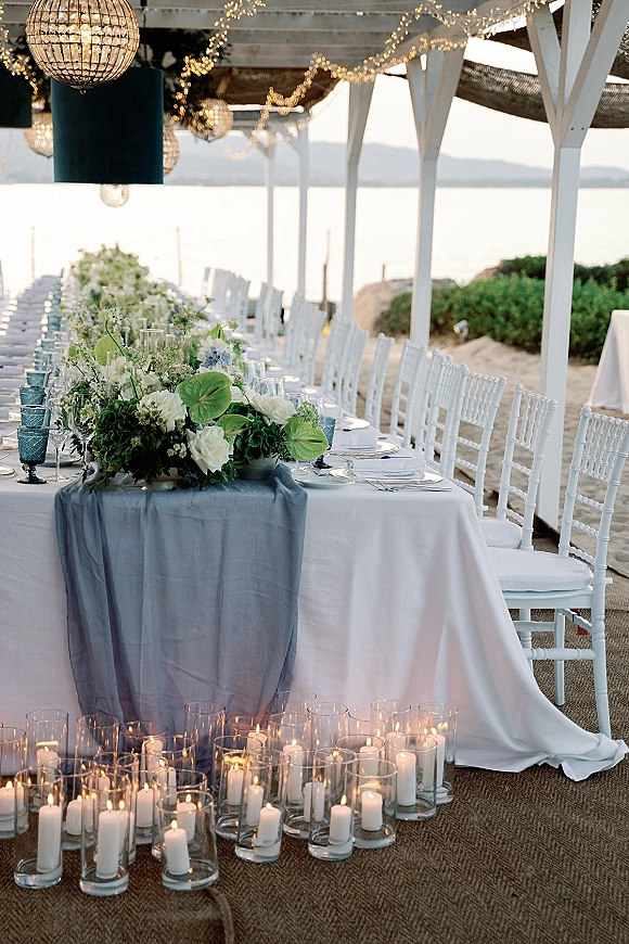 Reception tablescape on a long banquet table with blue runner, floral and candle centerpiece, blue goblets, and string lights by the waterfront