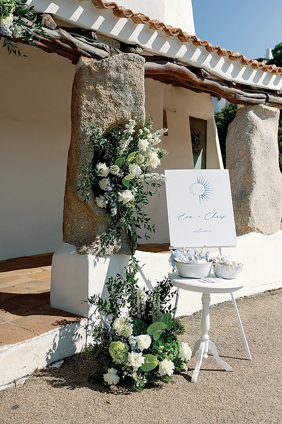 Wedding welcome sign on a wedding sign on easel beside white roses and greenery on a pedestal table, set by stone columns in sunlit patio entryway