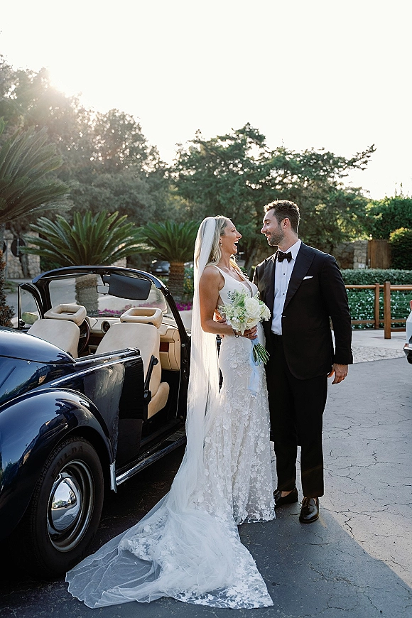 Couple portrait of bride and groom laughing beside a vintage convertible car, her long veil flowing, bouquet in hand on a sunlit driveway with palms