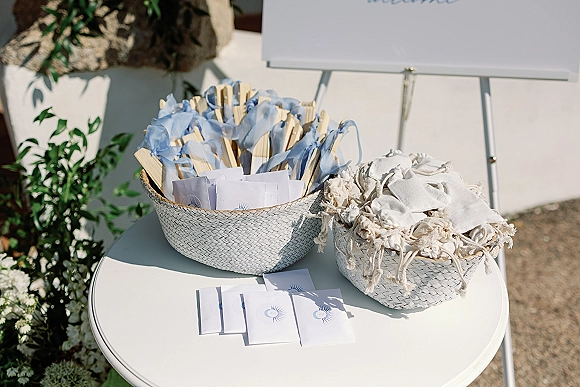 Wedding ceremony favors arranged in woven baskets with blue ribbon hand fans, paper programs, and muslin bags on a white table outdoors
