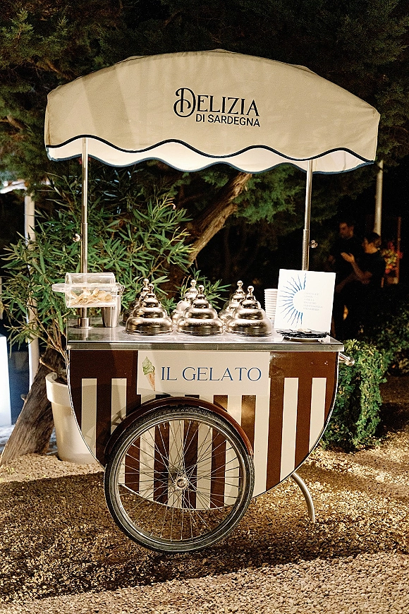 Gelato cart with a striped canopy, metal serving domes, cones, and menu card set on gravel in an outdoor garden reception area