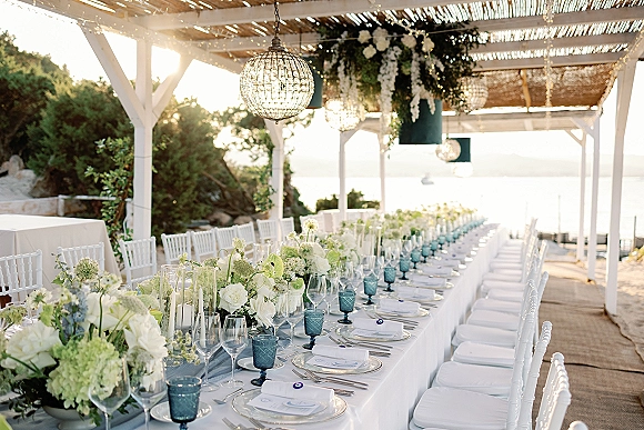 Reception tablescape with a long banquet table wedding setup, white florals, greenery garland, blue goblets, candle hurricanes under a pergola by the water