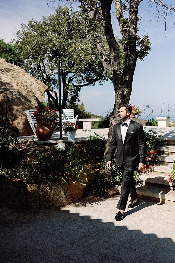 Groom portrait in a black tuxedo with bow tie and pocket square, walking on stone terrace steps with potted flowers and blue sky