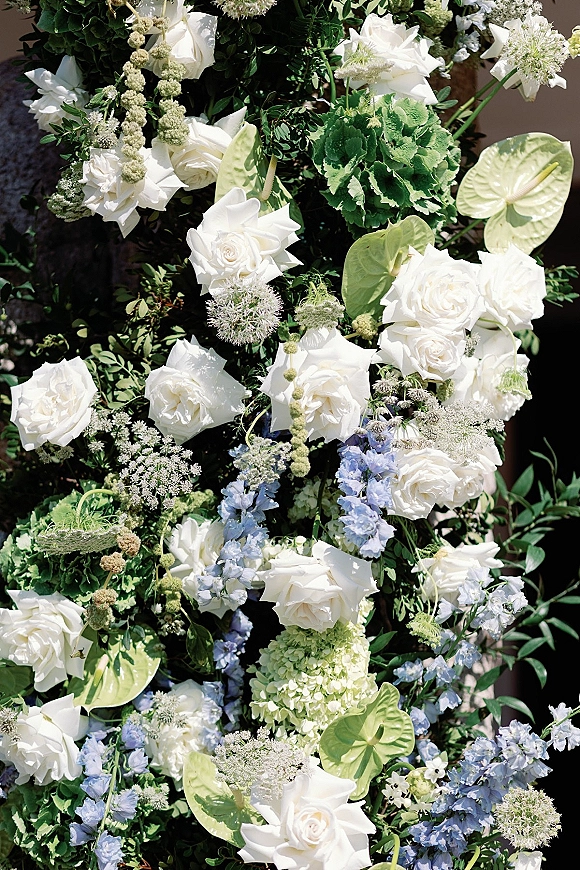 Wedding floral installation with white roses and blue delphinium, lush greenery and hanging amaranthus against a neutral wall in outdoor light