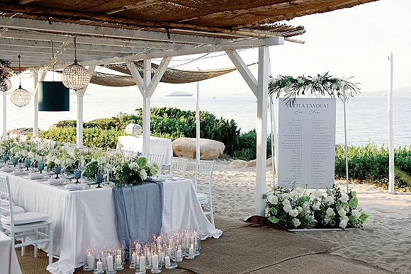 Beach wedding reception with long banquet table, blue runner, white chairs, florals, candles and string lights under a fabric canopy by the ocean.