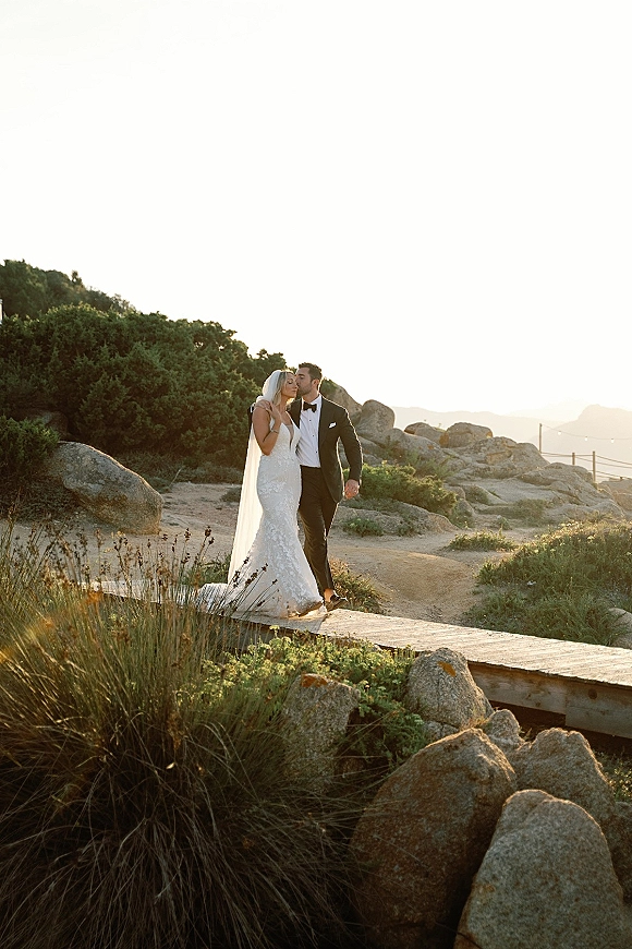 Wedding couple portrait of groom kissing bride’s forehead as her long veil flows, standing on a coastal boardwalk by boulders and string lights