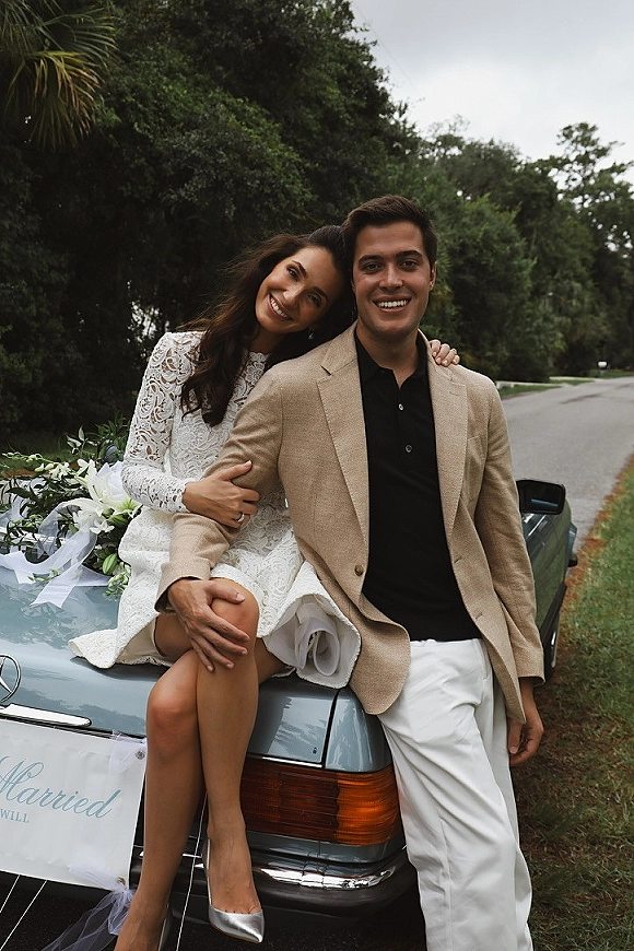 Couple portrait beside a wedding car photo, bride on the hood and groom leaning in front of a just married sign on a tree-lined road