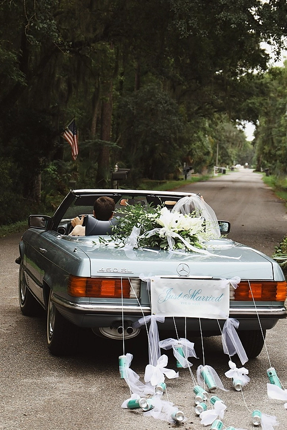 Just married car with wedding getaway car decor, white ribbons and tin cans trailing, floral greenery on trunk along a tree-lined road