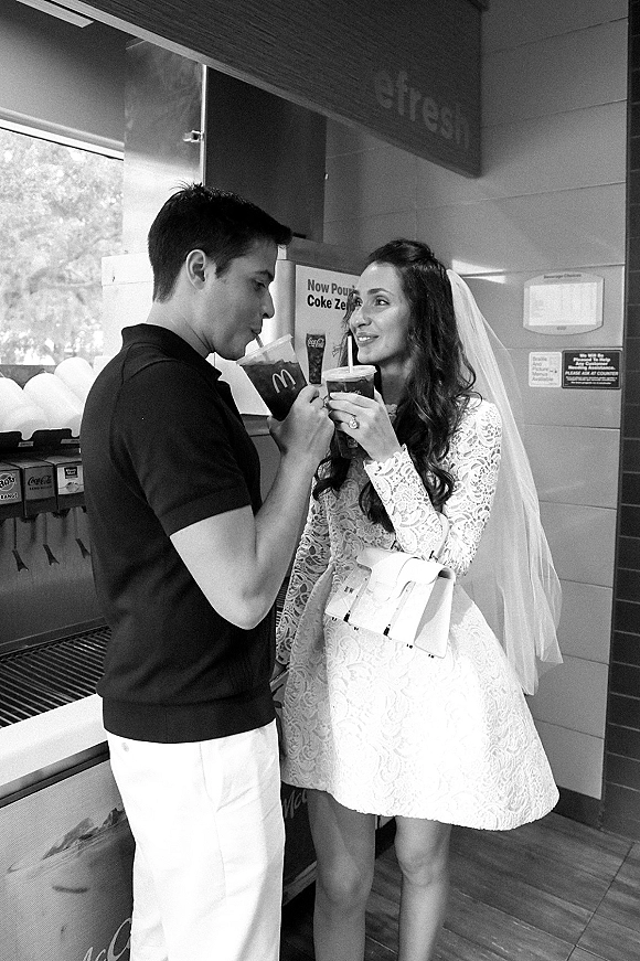 Couple portrait of bride in a short lace dress and veil and groom sipping soda in a fast food restaurant with menu signage behind them
