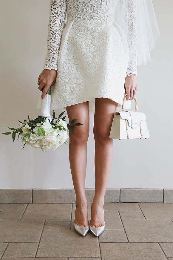 Wedding dress with short wedding dress lace long sleeves, veil, white rose bouquet, handbag and metallic heels on a tiled floor by a neutral wall