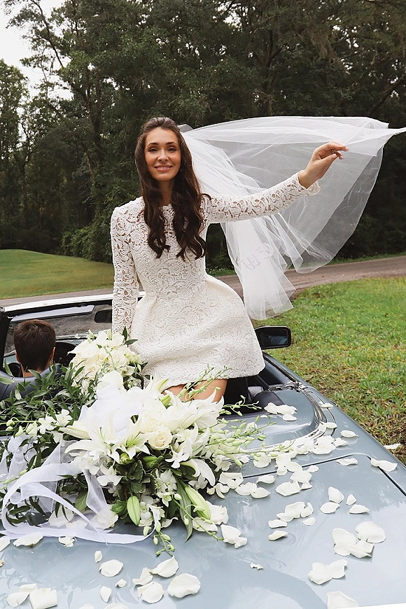Bride portrait in a lace long sleeve wedding dress, holding her veil and a white lily bouquet on a convertible with rose petals on the hood