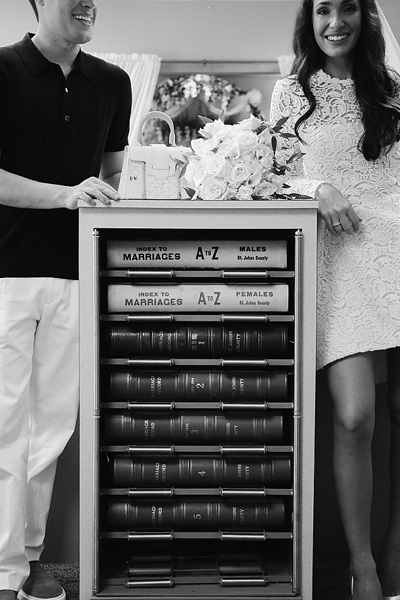 Couple portrait in a wedding courthouse portrait setting, bride in a lace mini wedding dress holding bouquet beside groom by records cabinet