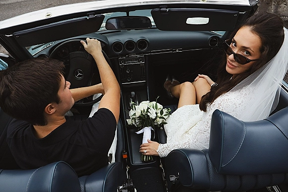 Newlywed car portrait of a bride in convertible looking back in sunglasses, veil trailing, holding a bouquet on leather seats