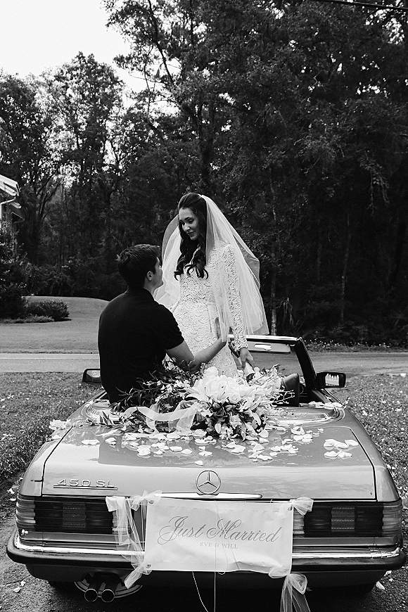 Wedding couple portrait of newlyweds on car, bride in a long veil beside groom on a decorated convertible on a tree-lined driveway