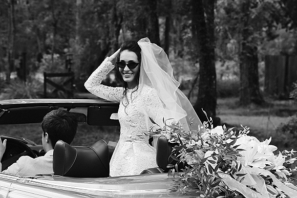 Wedding couple in a convertible getaway car, bride in sunglasses and lace dress with veil holding a bouquet, trees behind
