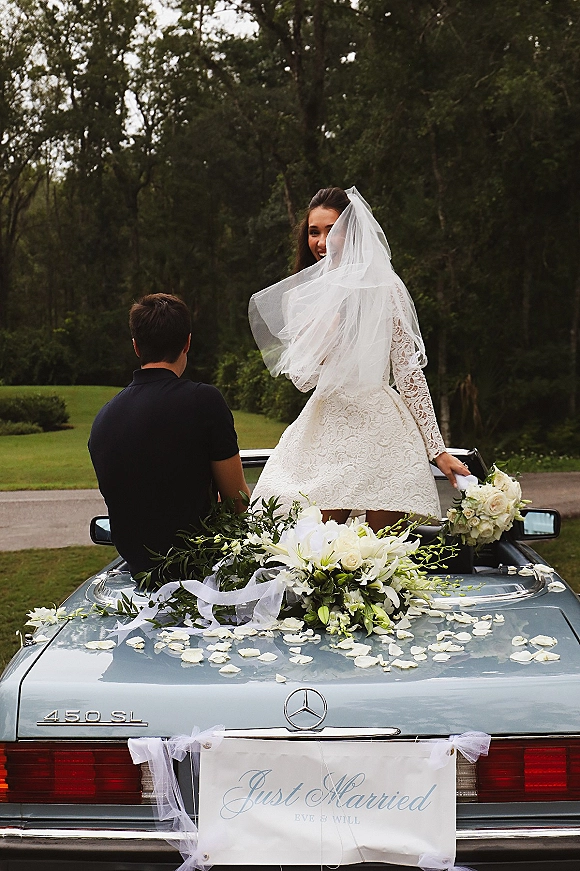 Wedding getaway car with a just married car sign, white ribbon bows and roses as the bride in lace sits in a convertible on a tree-lined road