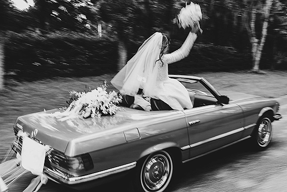 Wedding getaway car with bride in convertible holding a bouquet, veil blowing behind as the decorated car drives past trees and hedges